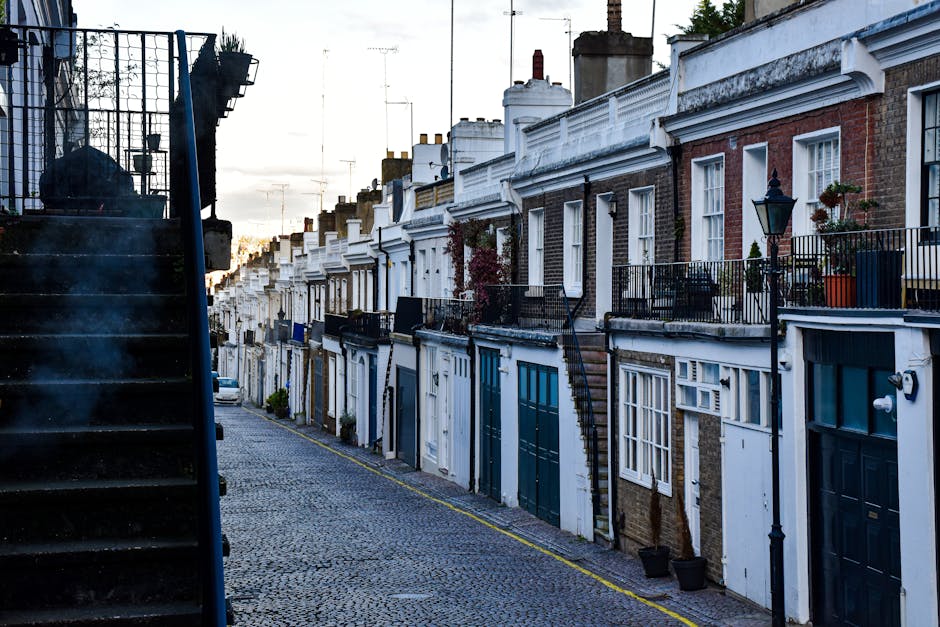 A narrow residential street in North Kensington shown during daytime, featuring a row of terraced houses with brick and white-painted facades, each with small front gardens or balconies decorated with potted plants and outdoor furniture. The street is paved with cobblestones and lined with black metal railings, while a black metal staircase on the left side leads up to an elevated entrance. Some houses have external staircase access to upper floors, and a few have small bay windows. The scene includes street lighting attached to the houses and a generally overcast sky overhead, indicating typical urban logistics for house removals and furniture transport. This environment reflects the typical challenges experienced during home relocation and packing and moving activities handled by companies such as Man with Van North Kensington, focusing on loading processes and moving logistics in historic residential areas.