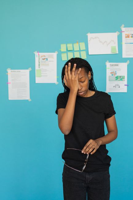 A young woman with dark braided hair, wearing a black t-shirt, stands indoors against a bright blue wall decorated with various printed sheets, charts, and sticky notes taped to the wall. She appears to be experiencing frustration or stress, with her right hand placed on her forehead and her eyes closed. The environment suggests an office or workspace setting related to house removals or moving logistics, with the presence of informational materials on the wall. The woman's posture and expression convey a sense of difficulty or concern, possibly related to the complexities of home relocation or packing challenges, which are relevant to the services provided by Man with Van North Kensington. The scene emphasizes the importance of professional support when facing problems during the moving process, such as access issues or logistical obstacles.