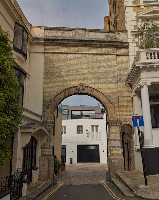 A narrow street in North Kensington featuring a historic stone archway connecting two brick buildings, with a modern white building with large windows visible through the arch. The street is paved with cobblestones and has yellow parking restriction lines along the curb. On the right, there is a white columned building with decorative architectural details, including a balcony. A blue 'no through road' traffic sign is mounted on a pole next to the white building. The scene is outdoors during daylight, with natural lighting illuminating the buildings and street. Man with Van North Kensington's team may use such narrow streets and architectural features when planning home relocation or furniture transport, involving careful logistics and precise loading and unloading processes.