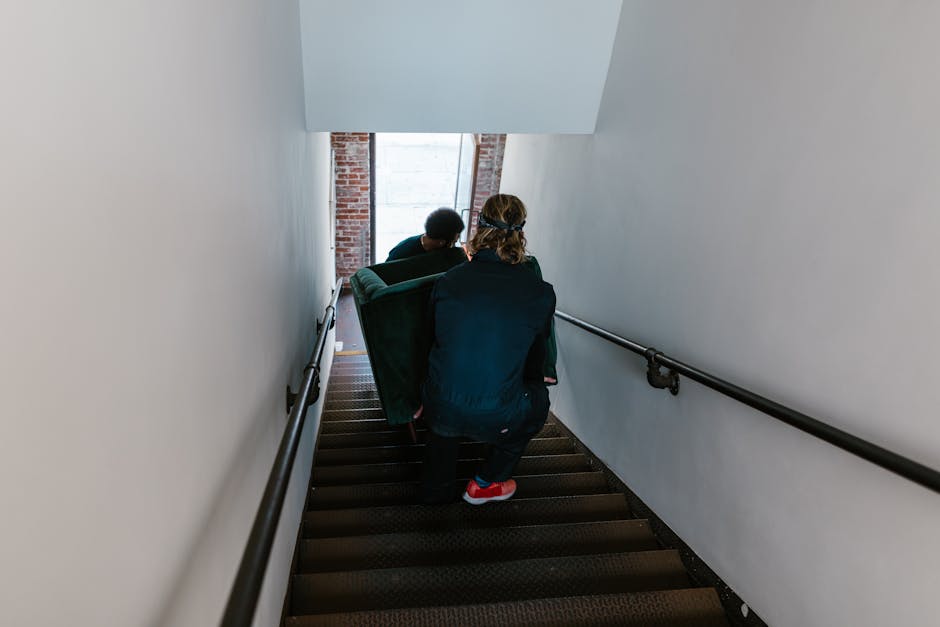 A man and a woman standing in a residential hallway beside a staircase, both holding cardboard boxes related to house removals. The man, dressed in a maroon shirt and orange trousers, is holding a lar