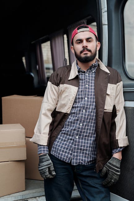 A man with a beard and wearing a backwards red cap, a checkered shirt under a color-blocked brown and beige jacket, and grey work gloves, standing inside a van with open side door during a home relocation. Behind him, the van's interior shows cardboard boxes stacked and ready for loading or unloading. The setting appears to be outdoors, with natural light illuminating the scene, capturing the process of furniture transport and packing for a house move. This image is relevant to professional removals services provided by Man with Van North Kensington, highlighting the loading process and logistical aspects of a house removal in the North Kensington area.