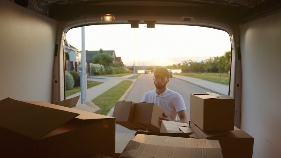 A man wearing a white shirt and a cap is standing outside the back of a moving van during a home relocation, with the van's open rear door revealing the interior filled with cardboard boxes of various sizes, some partially opened and others sealed, placed on the van floor. The man is holding a medium-sized box, preparing to load or unload it, as the setting sun casts warm light over a suburban street with houses, greenery, and a sidewalk visible in the background. The scene depicts the packing and loading process typical of furniture transport and house removals, with the focus on careful handling of packed boxes as part of a professional moving service provided by Man with Van North Kensington, supporting tight access moves around Ladbroke Grove and North Kensington.