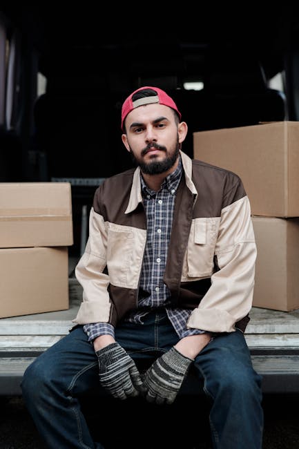 A man with a beard and wearing a backwards red cap, a checkered shirt under a color-blocked brown and beige jacket, and grey work gloves, standing inside a van with open side door during a home relocation. Behind him, the van's interior shows cardboard boxes stacked and ready for loading or unloading. The setting appears to be outdoors, with natural light illuminating the scene, capturing the process of furniture transport and packing for a house move. This image is relevant to professional removals services provided by Man with Van North Kensington, highlighting the loading process and logistical aspects of a house removal in the North Kensington area.