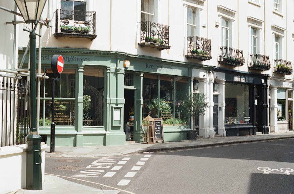 An exterior view of a row of commercial buildings on a quiet street in North Kensington, with a focus on a shop featuring large glass windows displaying plants and flowers. The building has a light green facade with decorative molding and a small awning, with a blackboard sign outside advertising flower arrangements. Adjacent to this shop is another storefront with a black entrance, possibly an art gallery or similar business. Above the shops, there are three floors of residential apartments with white walls and black wrought-iron balconies decorated with potted plants. The street has a designated bike lane marked with white paint and a pedestrian crossing with dashed lines, while a street sign and lamp post are visible on the pavement. The overall scene suggests a typical London neighbourhood with mixed-use buildings and a calm, inviting atmosphere, with no visible moving activity or furniture in the image.