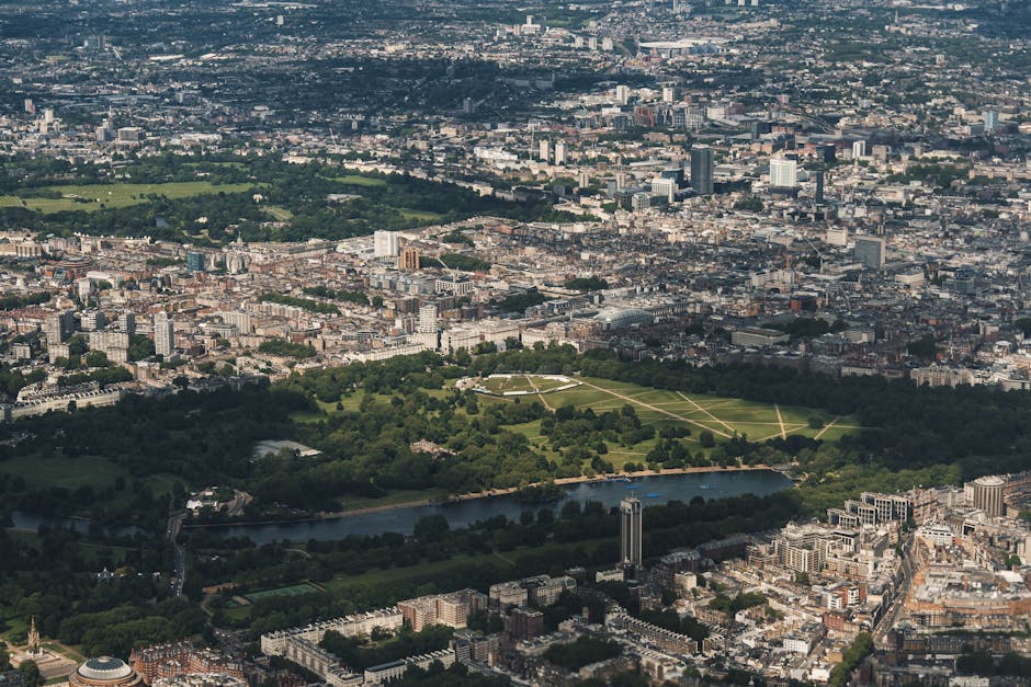 An aerial view of a large urban park surrounded by a densely built cityscape in North Kensington, London. The park features expansive green lawns, tree-lined pathways forming a geometric pattern, and a winding river running along its edge. In the foreground, there are residential buildings and streets, with a tall building near the river. The scene captures an urban home relocation setting, with natural daylight illuminating the area, providing context for house removals and furniture transport activities occurring nearby, as seen through the general aerial perspective. Man with Van North Kensington’s services relate to this environment, supporting efficient packing, loading, and home removals within this city context.