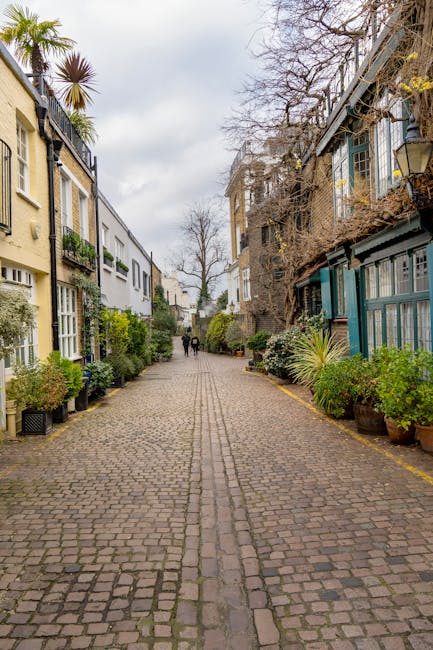 This image shows a cobblestone street in North Kensington, lined with residential buildings and lush potted plants along the edges. The buildings feature a mix of architectural styles, some with traditional bay windows and others with modern design, and are set against a cloudy sky. In the distance, a group of people is walking along the sidewalk. The scene captures an urban neighbourhood with a calm atmosphere, ideal for home relocation projects. Although no furniture or boxes are visible, the street's setting reflects the typical environment where Man with Van North Kensington offers same-day removals and furniture transport services, involving packing, loading, and moving household items within this residential area.