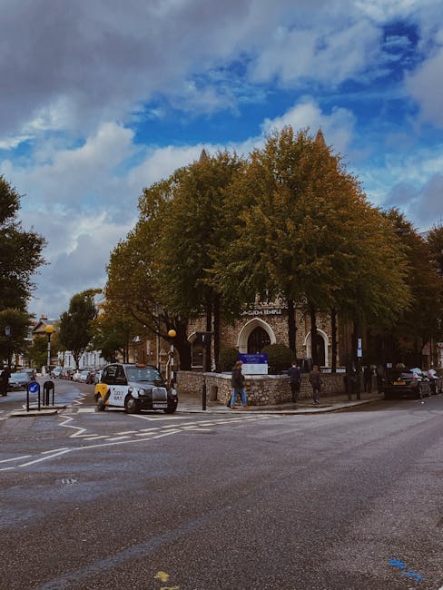 A street view featuring a small stone building with an arched entrance, surrounded by large trees with green and brown leaves indicating autumn, under a partly cloudy sky. In front of the building, a paved area is visible with a few parked cars and pedestrians walking past, some of whom are carrying or pushing moving boxes and small items. A man with a trolley is visible near the pavement, possibly involved in loading or unloading goods for a house removal or relocation service. The scene captures a typical urban setting suitable for house removals in North Kensington, with a focus on moving logistics such as furniture transport and packing within the context of a home relocation. Man with Van North Kensington provides professional removals, evidenced by the structured environment and activity around the property.
